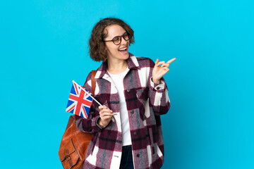 Young English woman holding an United Kingdom flag isolated on blue background pointing finger to the side and presenting a product