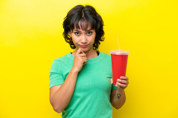 Young Argentinian woman holding a soda isolated on yellow background thinking