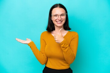 Fototapeta premium Young caucasian woman isolated on blue background holding copyspace imaginary on the palm to insert an ad and with thumbs up