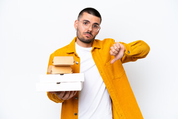 Young caucasian man holding pizzas and burgers isolated on background showing thumb down with negative expression