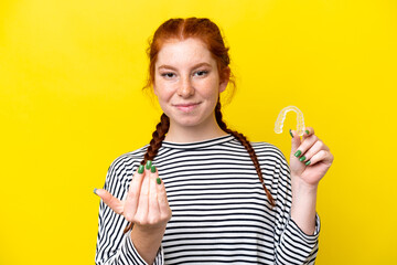 Young caucasian reddish woman holding invisible braces isolated on yellow background inviting to come with hand. Happy that you came