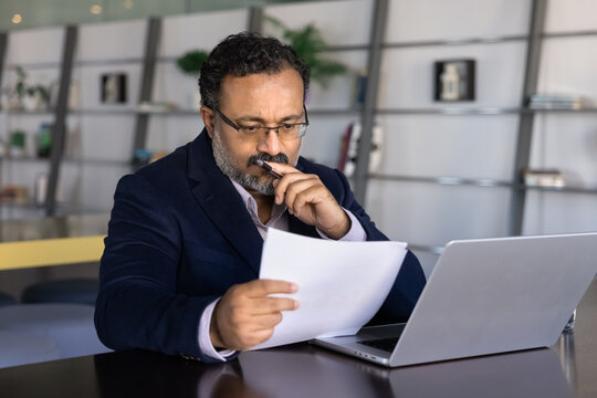 Thoughtful frowning senior Indian businessman reading paper document at laptop, working at computer in co-working office, reviewing invoice for tax payment, touching chin