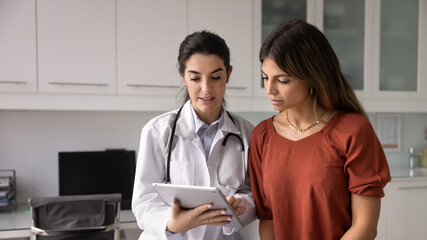 Optimistic young Hispanic doctor woman showing medical report on tablet to female patient sitting on couch, telling checkup result, giving prescription, treatment advice. Banner shot © fizkes