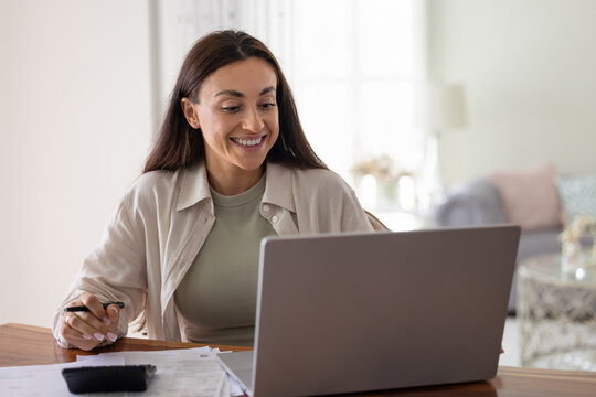 Young bookkeeper woman working attentively from home office, sit at table with laptop, using digital tools and printed documents. Small business management, entrepreneurship, personal budget planning