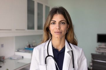 Serious friendly beautiful Hispanic female doctor wearing lab coat, standing in clinic examination room, posing for head shot, looking at camera. Medical healthcare professional woman portrait