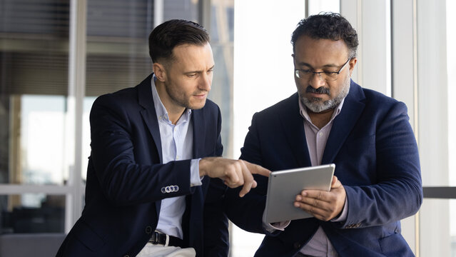 Two serious different aged diverse male coworkers using digital tablet together, reviewing online product presentation. Elder manager consulting younger colleague, showing gadget to expert