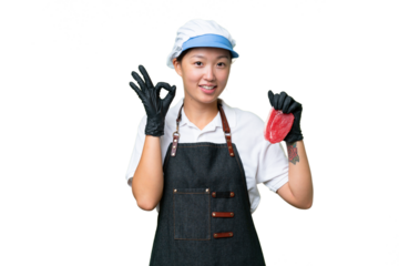 Young caucasian woman wearing an apron and serving fresh cut meat over isolated background showing ok sign with fingers