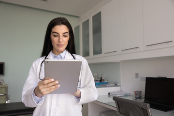 Focused young Latin doctor woman browsing electronic medical records on tablet, using modern online technology, chatting with patients, giving support, consultation on Internet