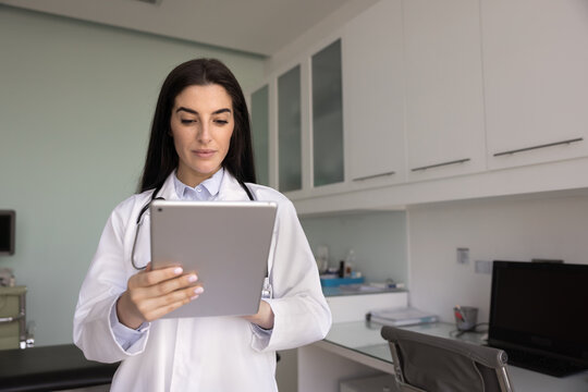 Focused young Latin doctor woman browsing electronic medical records on tablet, using modern online technology, chatting with patients, giving support, consultation on Internet - Powered by Adobe