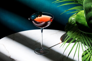 Strong alcoholic cocktail drink with whiskey, vermouth, bitters and blue cherry. Marble table, stylish background with green plants