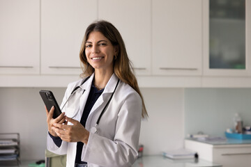 Happy successful female Hispanic doctor woman holding mobile phone, posing I hospital office for medical professional portrait, looking at camera, smiling, using gadget for online consultation