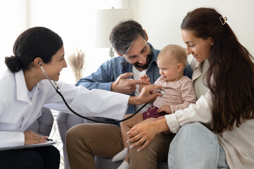 Smiling woman pediatrist in white coat using stethoscope to listen of baby heartbeat, cute infant sits on daddy lap smile during heart check-up. Professional healthcare services, paediatrics industry