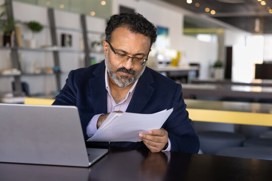 Serious focused elder Indian executive man reading document carefully, analyzing paper report at workplace with laptop, checking agreement text, bill, invoice for online payment