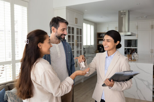Smiling Indian female, real estate agent hands over set of keys to married couple. Positive experience of finding new home using professional services of agency. Happy homeownership, new beginnings