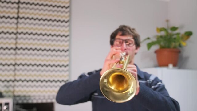 A man is captured in close-up during a home trumpet rehearsal, with puffed cheeks and intense focus. Facial expression and finger movement on the valves. A personal, intimate musical moment.