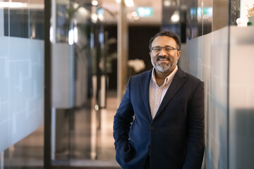 Cheerful senior Indian businessman posing in office hall with glass walls, looking at camera, laughing, keeping hand in pocket in confident gesture. CEO, executive business portrait with copy space
