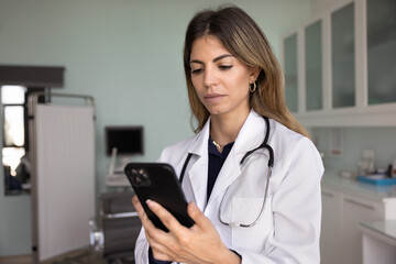 Serious female Hispanic doctor in white coat and stethoscope typing on mobile phone, using medical service for online communication and remote patients support from hospital office