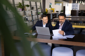Busy younger and elder multiethnic business coworkers working at laptop together in co-working space, discussing paper marketing reports, analyzing financial growth economic data