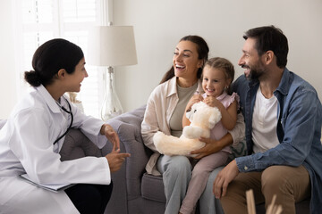 Friendly Indian pediatrician woman wearing white coat and stethoscope engaging in conversation with family and little 3s daughter sits on mommy lap holding favourite toy during therapist visit at home