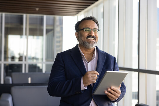 Cheerful happy older Indian business leader man using financial web service on tablet in office lobby, looking away, smiling, laughing, thinking on successful trading, income from investment