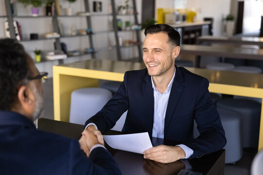 Happy younger Latin business company director man hiring senior job candidate after successful interview, holding paper resume, shaking hand of new employee, coworker, smiling