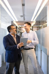 Fototapeta premium Cheerful diverse different aged business partners using tablet in office corridor, standing together, talking and laughing on work break, discussing creative ideas for online product. Vertical shot