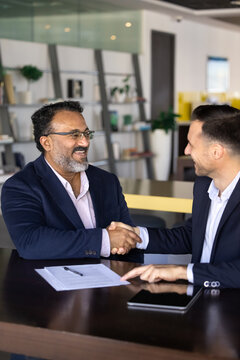 Happy business project buyer and seller men shaking hands, sitting at work table with documents, meeting for cooperation, partnership, getting agreement about investment. Vertical shot