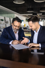 Positive older and young diverse business professional men signing contract at table. Consultant, agent, legal expert, financial advisor pointing at place for signature to customer. Vertical shot