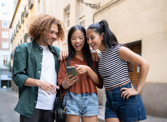 Three happy young tourists are using a smartphone and laughing while walking down a city street
