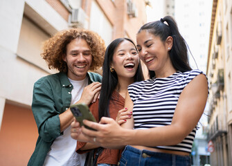 Three cheerful multi-ethnic friends are enjoying time together, laughing while looking at a smartphone in a city alley