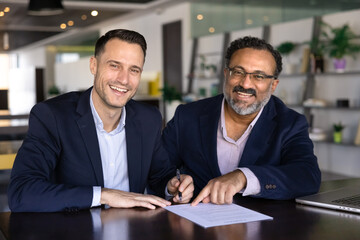 Two happy young and elder diverse male business partners getting agreement at work table, writing in document, signing paper agreement, contract, looking at camera, smiling, laughing