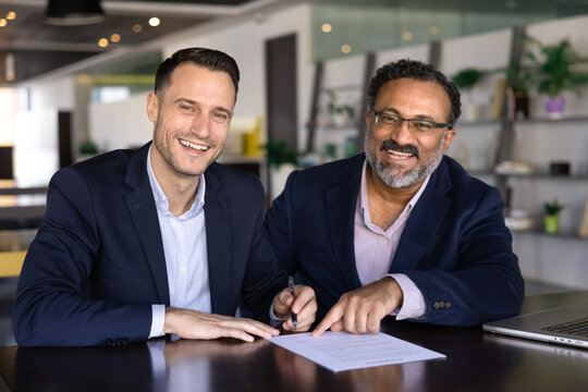 Two happy young and elder diverse male business partners getting agreement at work table, writing in document, signing paper agreement, contract, looking at camera, smiling, laughing