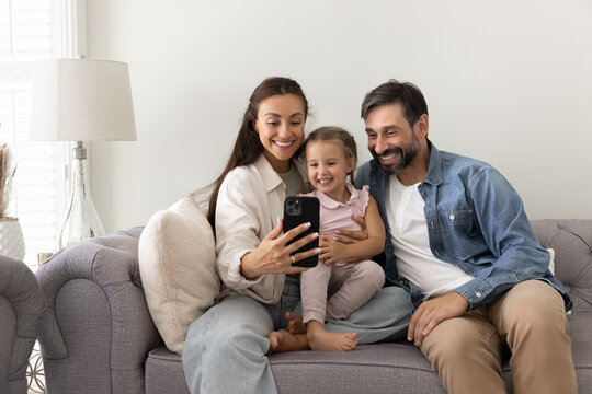 Family with little cute daughter using modern smartphone seated on cozy sofa in living room, making video call to relatives on holiday, take selfie picture, play new cool videogame on weekend at home - Powered by Adobe