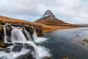 Moody view of Kirkjufell mountain in Iceland captured on an overcast day, with a long exposure smoothing the flowing waterfalls and the still waters of the lake. The dramatic light and soft textures c