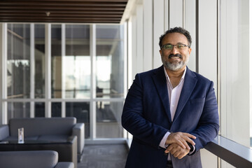 Happy successful older Indian business leader man in glasses and formal clothes posing in office lobby space alone, looking at camera, smiling. Successful mature CEO, executive professional portrait