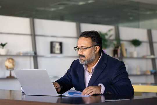 Serious busy elder Indian executive man using online business application on computer for marketing analysis, checking Internet sales statistics, typing on laptop for communication