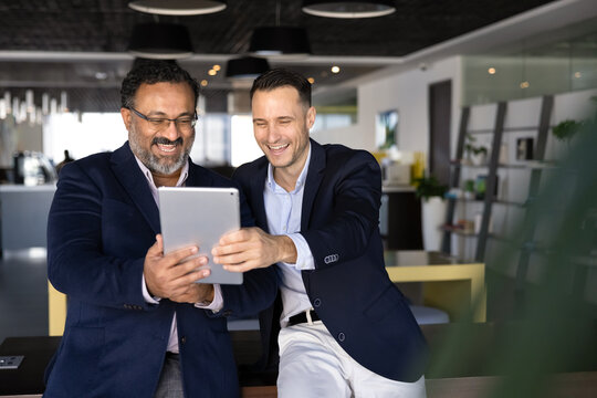 Two cheerful excited younger and elder diverse office colleagues using digital tablet together for fun, taking selfie, talking on online video call, enjoying Internet communication, smiling, laughing