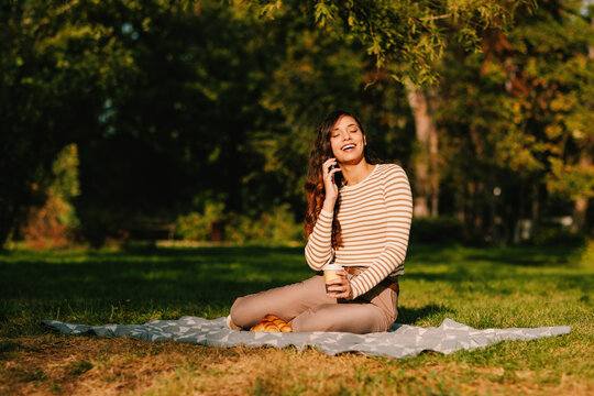 Young woman enjoying a relaxing picnic in the park while talking on her phone - Powered by Adobe