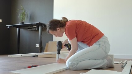 Woman using cordless drill to screw part into white bed frame. Female assembling furniture in new apartment. DIY home renovation and home organization after moving concept