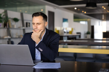 Fototapeta premium Serious young Latin businessman working at computer in co-working office space, using online professional service, managing Internet project, analyzing ebusiness startup, touching chin, thinking