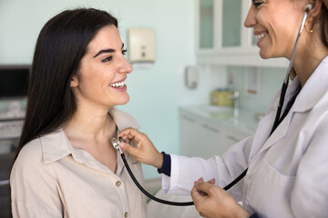 Happy female GP doctor examining positive young patient, listening to heartbeat and breathing, touching chest of woman with stethoscope, asking about healthcare, talking, smiling