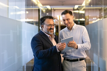 Positive multiethnic male younger and elder business colleagues using digital tablet in office corridor with glass walls, watching online product content on device, smiling, laughing