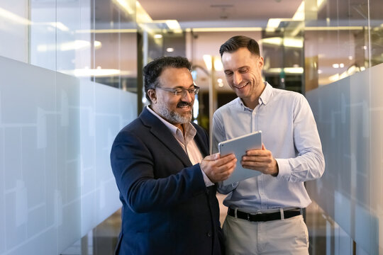 Positive multiethnic male younger and elder business colleagues using digital tablet in office corridor with glass walls, watching online product content on device, smiling, laughing - Powered by Adobe