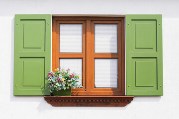 Wooden window with green shutters on a white wall. Decorative flower box with colorful blossoms below. Symmetrical architecture detail of traditional building. Bright daylight enhances vibrant colors.