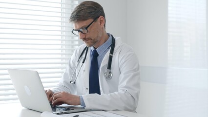 Concentrated male doctor in glasses and lab coat with stethoscope typing on laptop keyboard while sitting at table in medical office