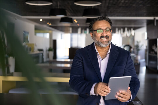 Cheerful elder Indian businessman using tablet computer, looking away, smiling, laughing. Senior executive holding gadget in co-working office, thinking on Internet technology for business success