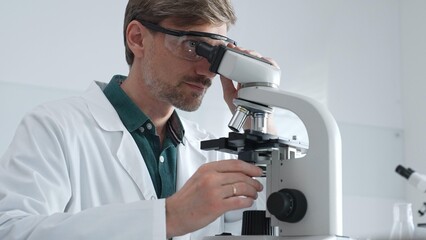 Adult male scientist wearing lab coat and protective glasses using microscope, doing scientific...