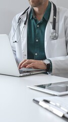 Doctor man wearing a white coat over green shirt, and stethoscope is typing on a laptop in medical office, showcasing the intersection of healthcare and technology, vertical close up view. Medicine