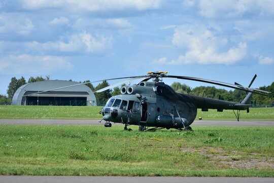 Large Mi-17 military transport helicopter on grassy airfield, front view with rotor blades extended, cockpit windows reflecting sunlight, hangar in background.