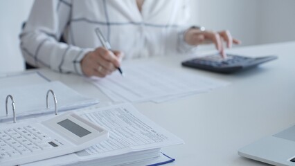 White calculator is on financial documents on the office desk, with an accountant taking notes, emphasizing accounting, finance, and tax preparation, close up. Audit and taxes concept